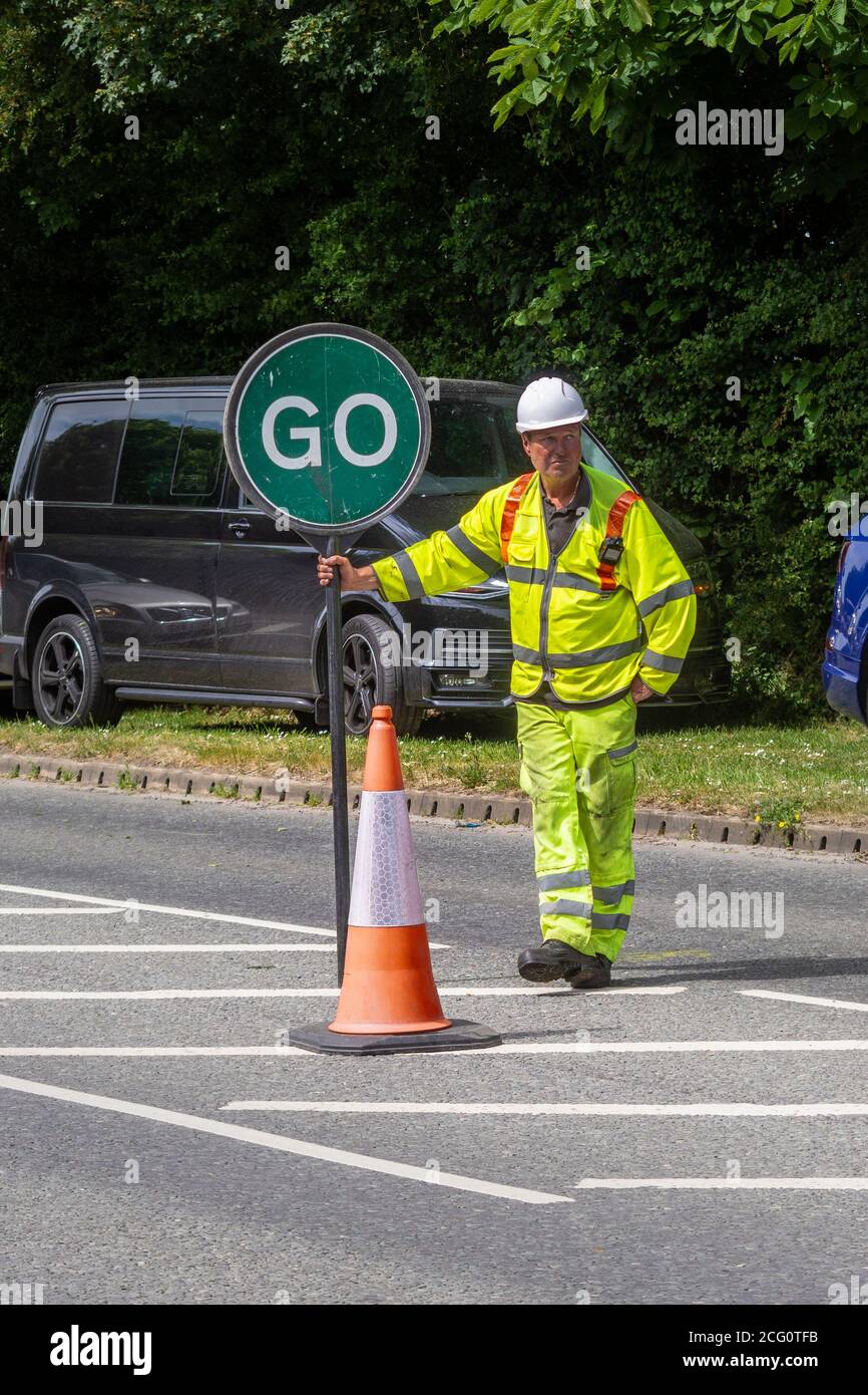 A traffic control worker in high visibility clothing with a traffic control lollipop with the green go side showing standing next to a cone in the roa Stock Photo - Alamy
